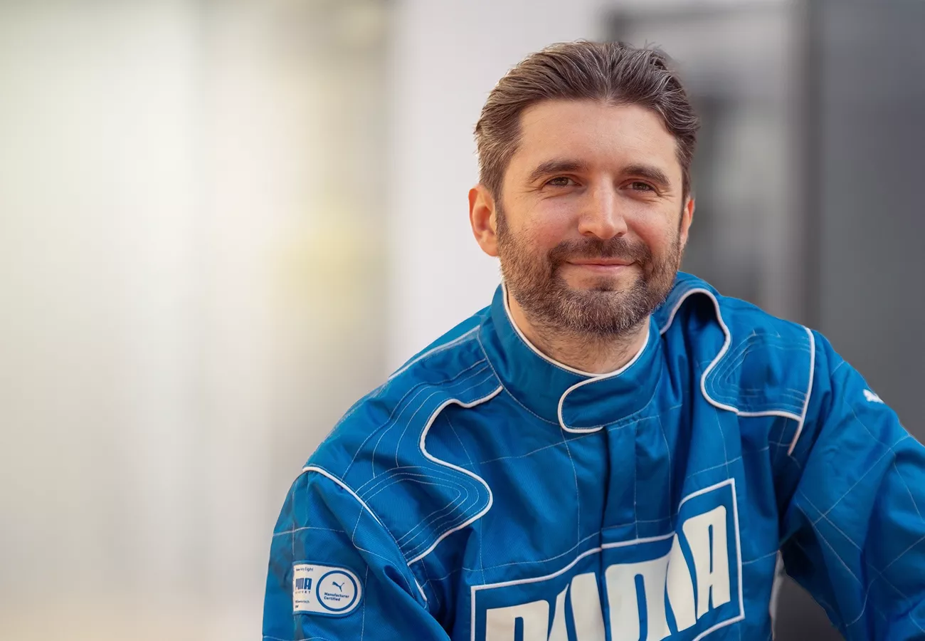 A person wearing a blue racing suit with visible white stitching and a bold logo is indoors. The setting appears modern and professional, with a blurred background. The suit features a patch with additional details on the sleeve.